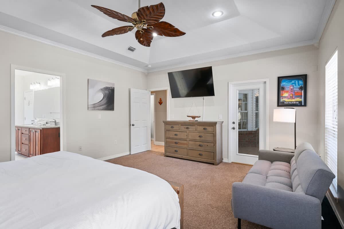 A wide shot of the master bedroom, highlighting the wooden dresser with a wall-mounted TV, a gray sofa, and an open doorway leading to the en-suite bathroom, all beneath a tray ceiling with a ceiling fan.