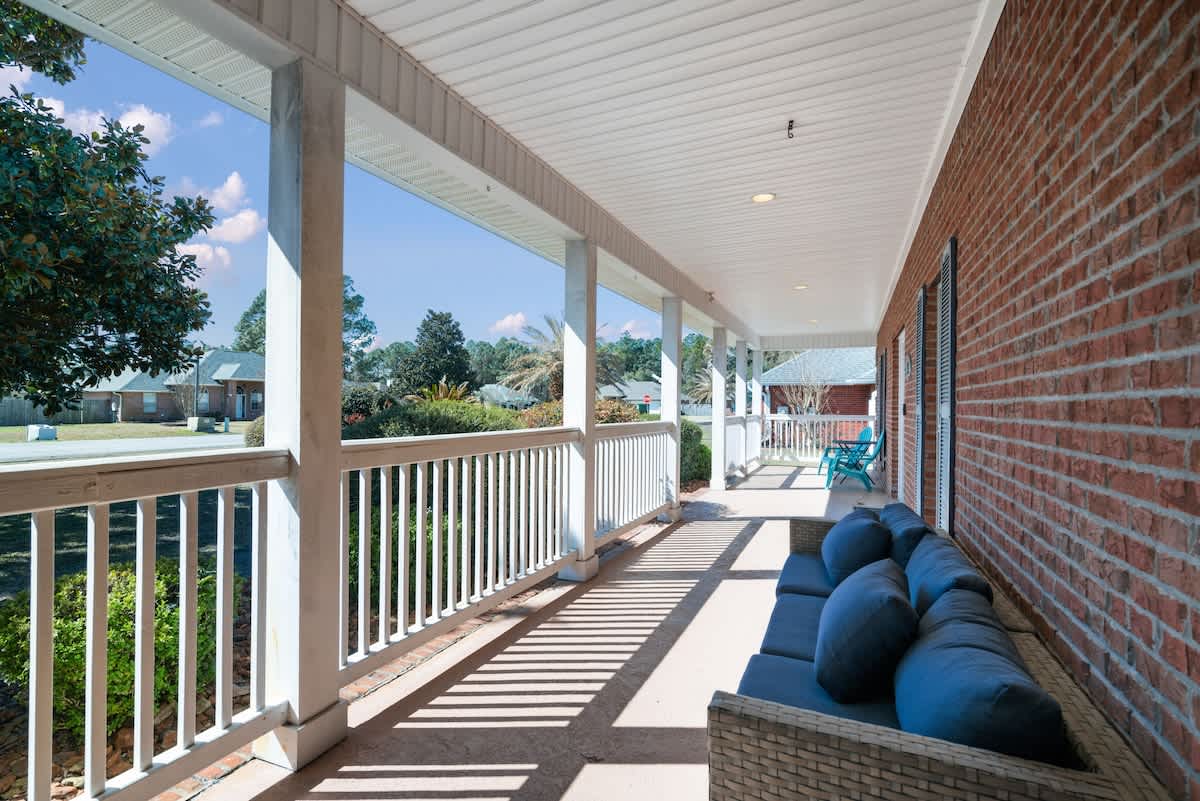 A wide-angle view of the covered front porch, featuring a red brick wall, white railings, and a wicker sofa with blue cushions, extending along the front of the house with ample space for relaxation.