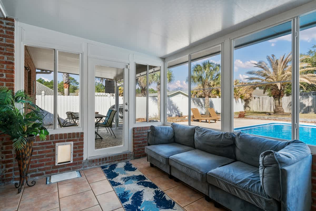 Comfortable seating area within the sunroom, featuring a large blue sectional sofa and a patterned rug on tiled flooring. A glass door leads to the outdoor patio, with large windows offering views of the pool and backyard.