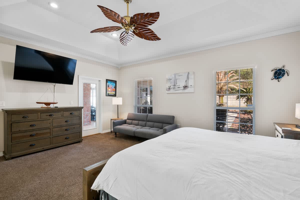 Another view of the master bedroom, showcasing a large wooden dresser with a wall-mounted TV above it, a gray sofa, and multiple windows with blinds, all under a decorative ceiling fan.