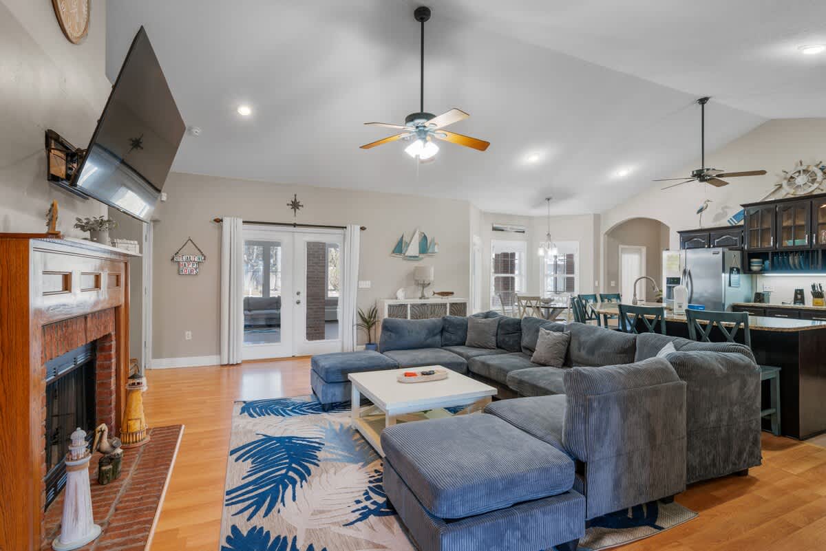 Living room with a blue sectional sofa and a white coffee table, featuring light wood floors and a vaulted ceiling. French doors open to an outdoor area, and a dining space is visible beyond.