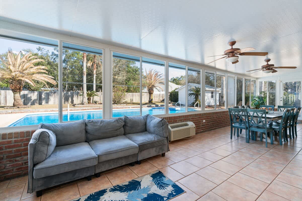 Bright and airy sunroom featuring a blue sectional sofa and a dining area with a large table, all set on terracotta-colored tiled flooring. Multiple ceiling fans and expansive windows provide views of the outdoor pool and palm trees.