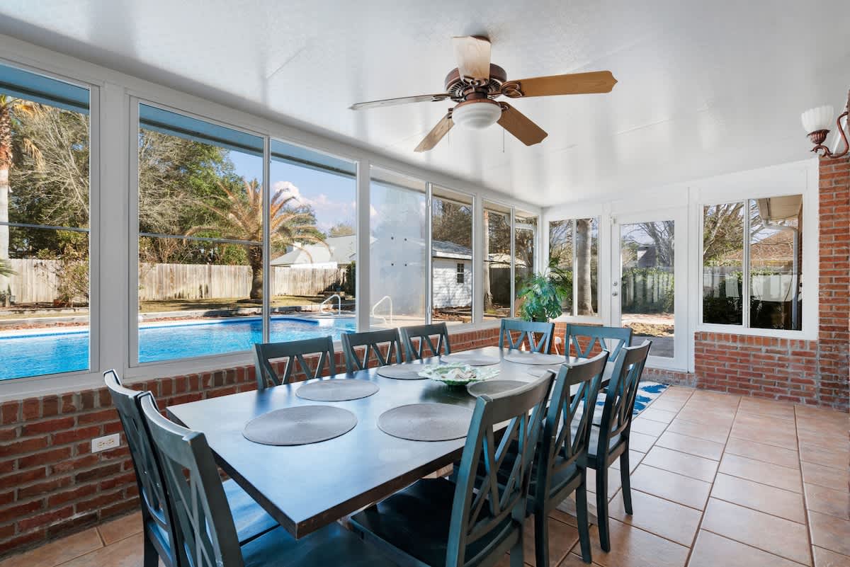 Bright sunroom serving as a dining area, featuring a large dark dining table with eight blue chairs on tiled flooring. Expansive windows offer panoramic views of the outdoor swimming pool and palm trees.