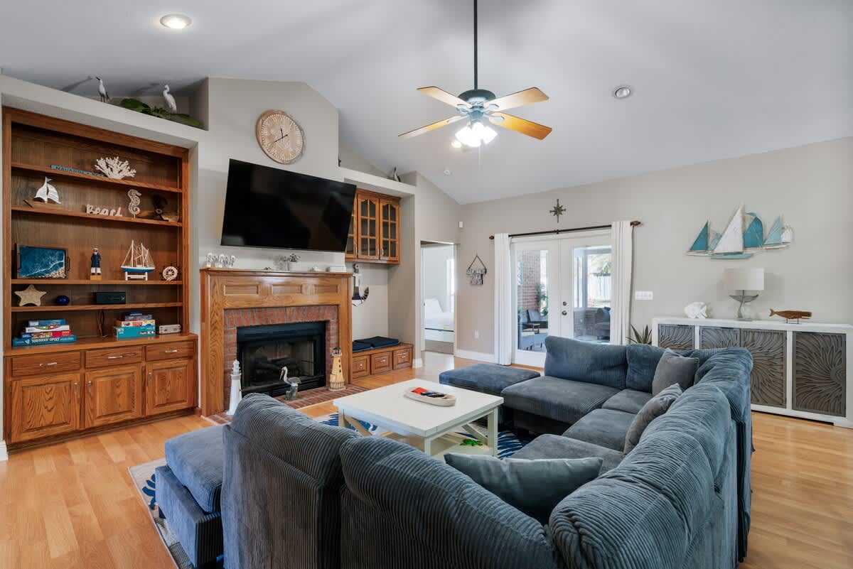 Spacious living room with a large blue sectional sofa, a brick fireplace with a mounted TV, and built-in wooden shelving filled with nautical decor. Light wood floors and vaulted ceilings complete the coastal-themed space.