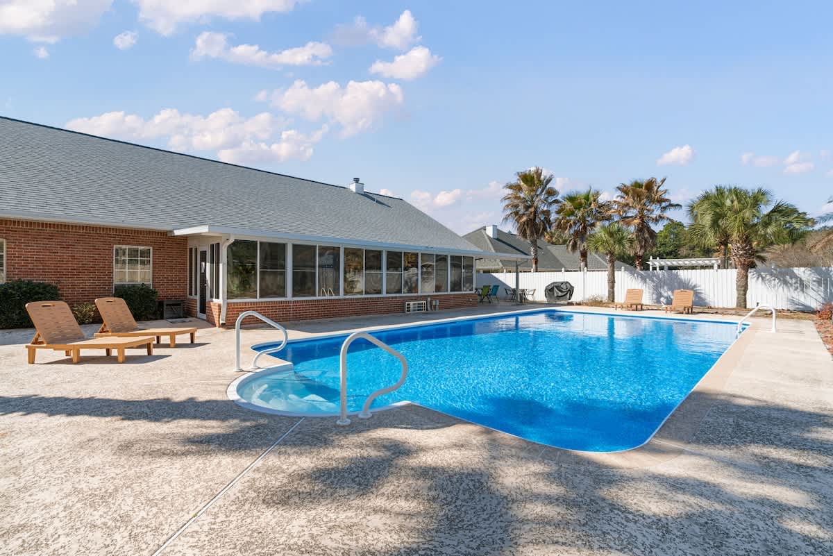 Wide shot of a large, bright blue outdoor swimming pool with a curved entry, surrounded by textured concrete decking. The brick house with a sunroom and several palm trees line the perimeter under a sunny sky.