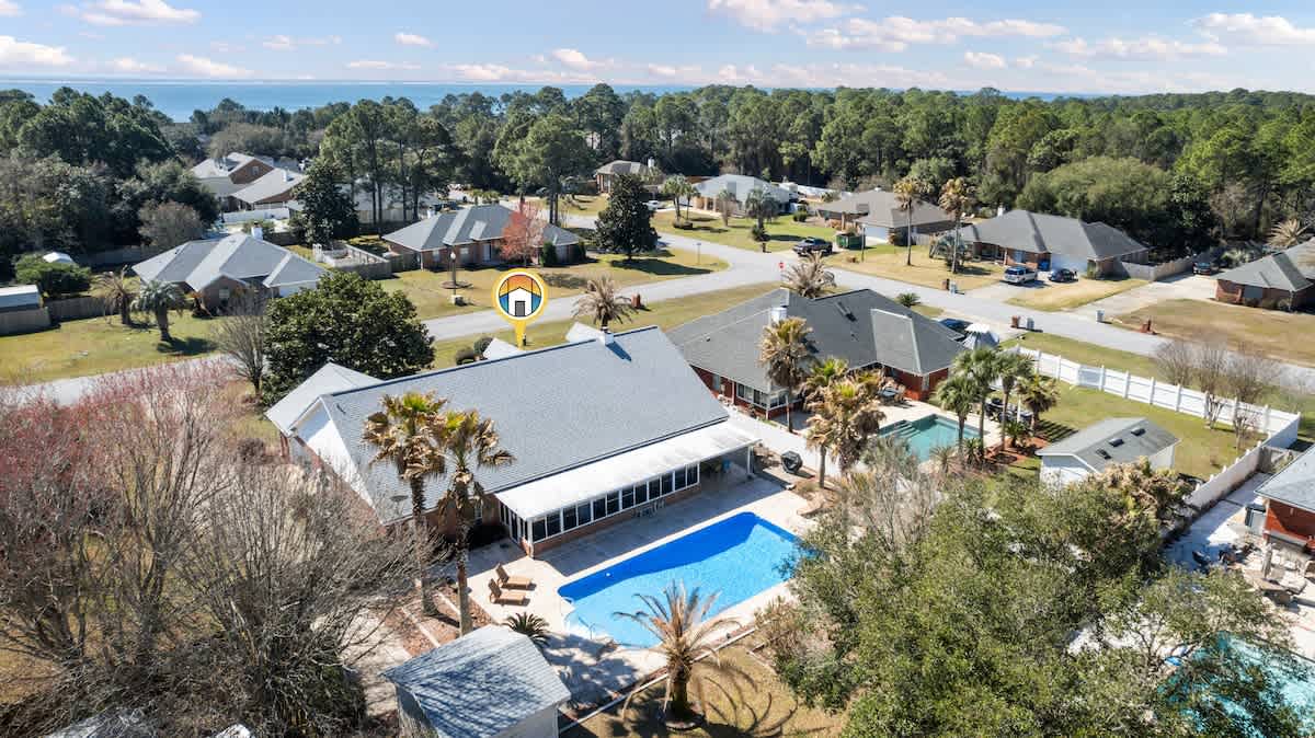 A slightly different aerial perspective of the house, highlighting the light gray roof and the backyard with a bright blue swimming pool, surrounded by mature trees and other residential homes, with the distant water still visible.