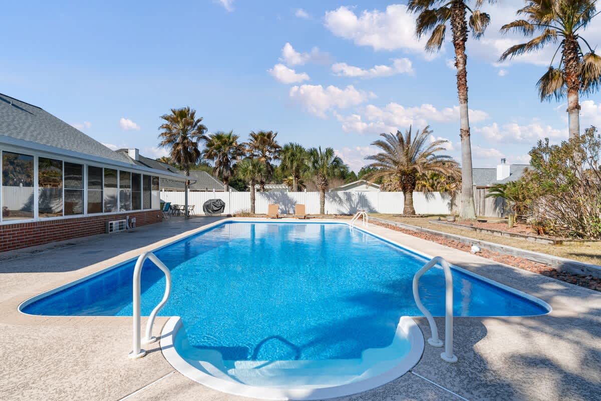 Inviting outdoor swimming pool with bright blue water, featuring white handrails and steps for easy entry. Several tall palm trees and a white fence are visible in the sunny backyard.