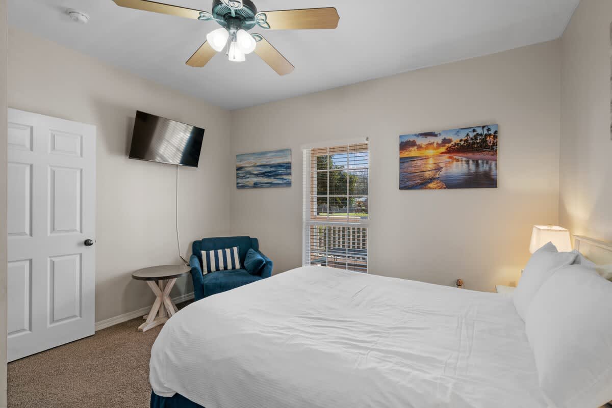 A bright bedroom with a white-covered bed, a blue armchair, and a wall-mounted TV. A large window with blinds and a beach-themed artwork above it provides natural light.