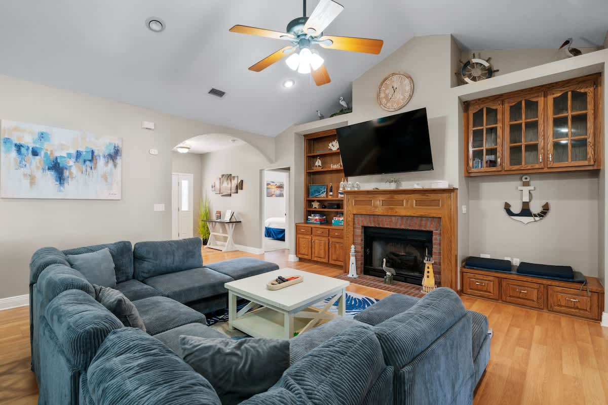 Wide-angle view of a coastal-themed living room featuring a large blue sectional sofa, a brick fireplace with a mounted TV, and light wood floors. An arched doorway leads to another part of the house.