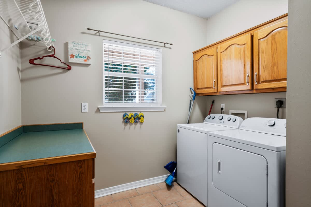A functional laundry room equipped with a white washer and dryer, wooden upper cabinets, a green countertop, a window with blinds, and wire shelving for storage, all on a tiled floor.