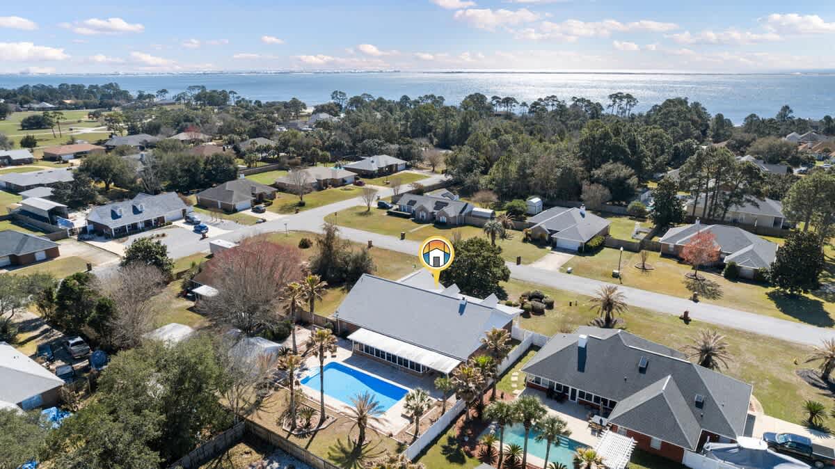 An aerial shot showcasing a residential property with a light gray roof and a bright blue swimming pool in the backyard, set within a green, tree-lined neighborhood with a large body of water in the background.