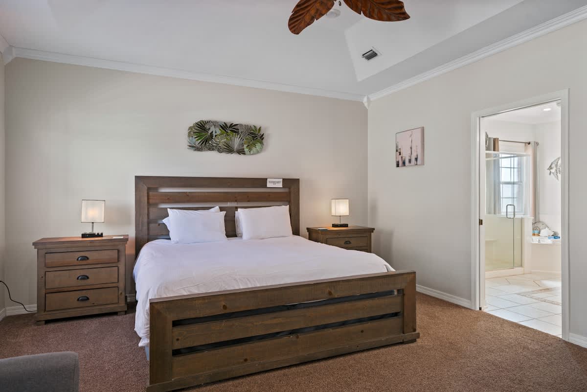 Close-up of the wooden king-sized bed with white bedding, flanked by two wooden nightstands with lamps, and an open doorway revealing the master bathroom beyond.