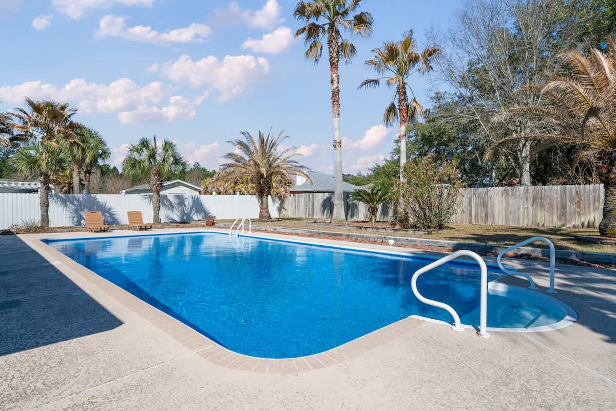 Spacious outdoor swimming pool with a bright blue liner and a curved entry, surrounded by light-colored concrete decking. The backyard features several mature palm trees and a mix of wooden and white fences.