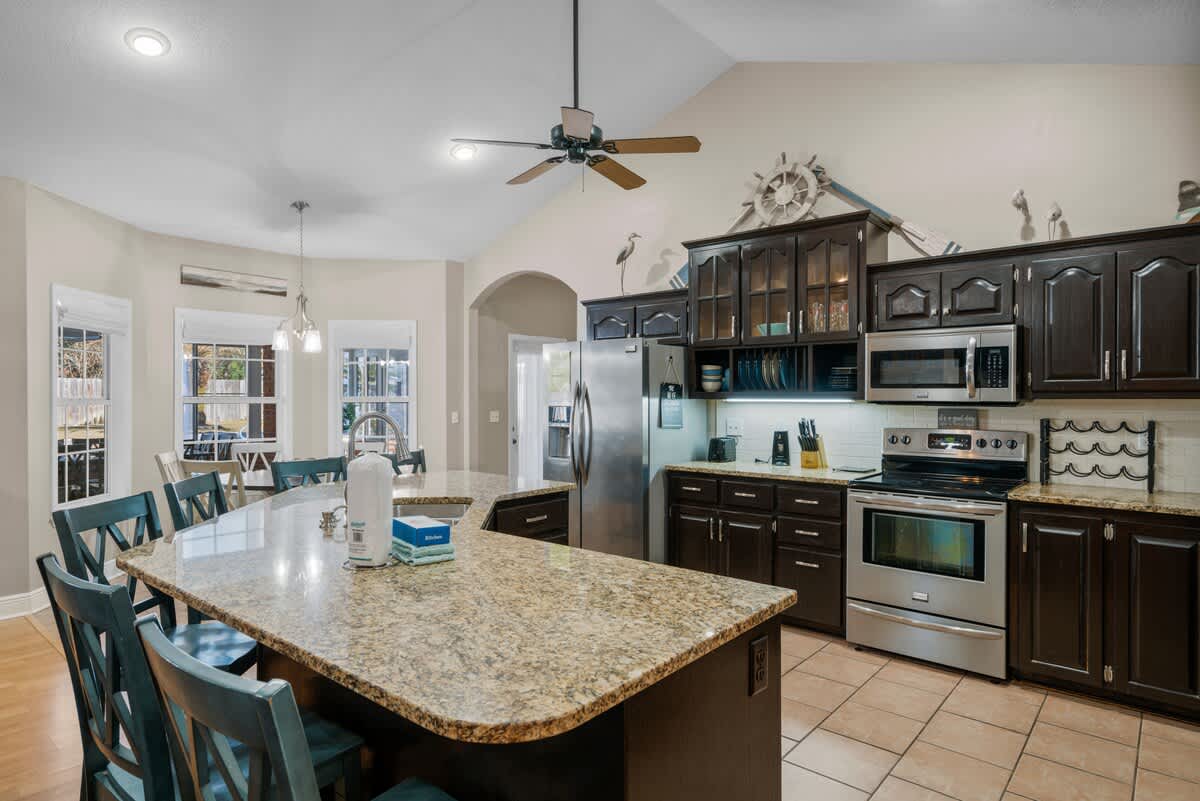 Spacious kitchen with a large granite island providing seating for four, dark wood cabinets, and stainless steel appliances. The area features light tile flooring and a ceiling fan.
