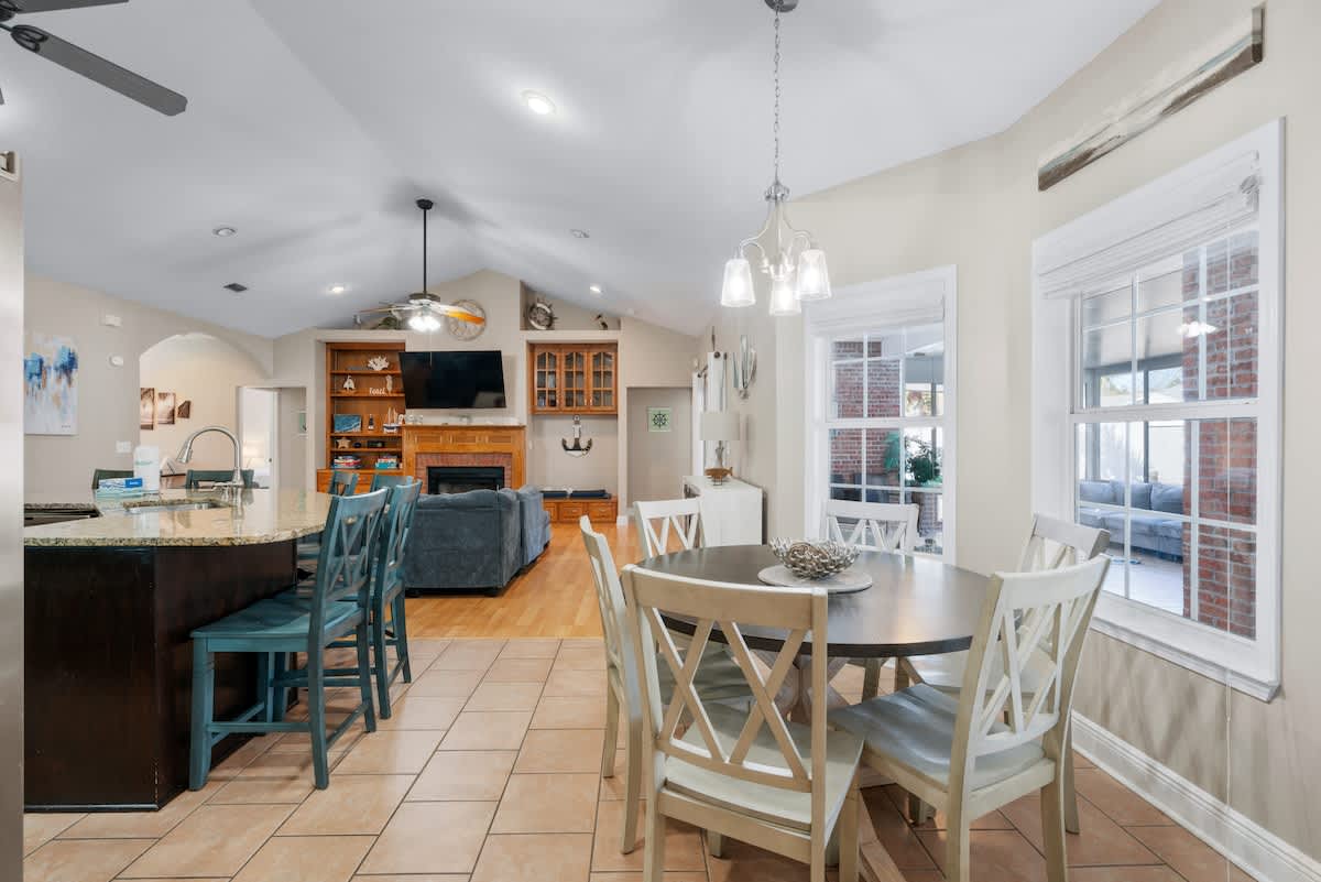 An open-concept dining area featuring a round wooden table with white chairs, a chandelier, and a tiled floor, with a view into the living room and kitchen island with bar stools.
