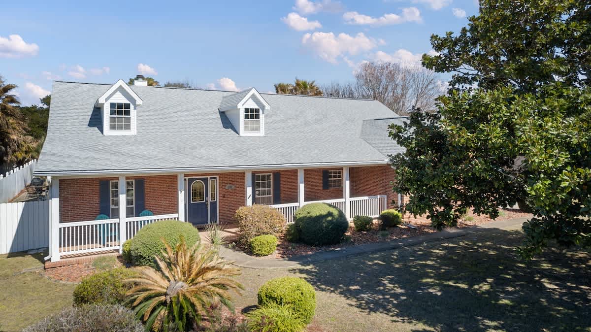A slightly closer view of the front of the brick house, showcasing the light gray roof, dormer windows, and the covered porch with white railings, surrounded by well-maintained green bushes and a large magnolia tree.