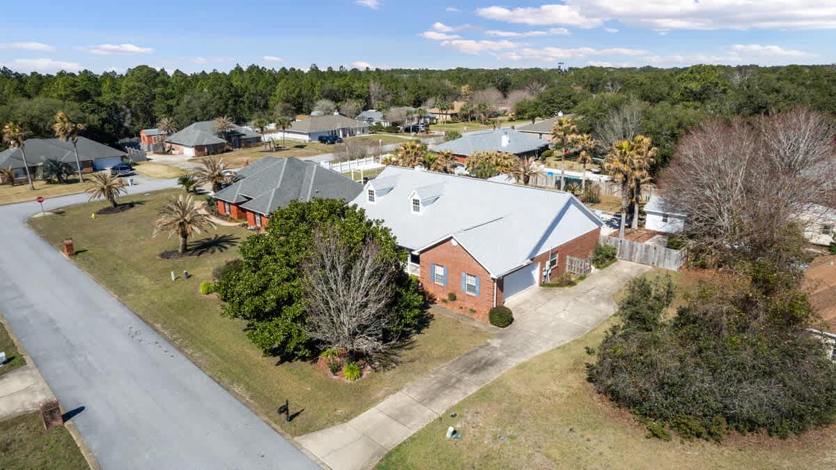 An aerial-like view of the front and side of the brick house with a light gray roof, showing the driveway leading to a garage, and a large mature tree in the front yard, with other houses visible in the background.