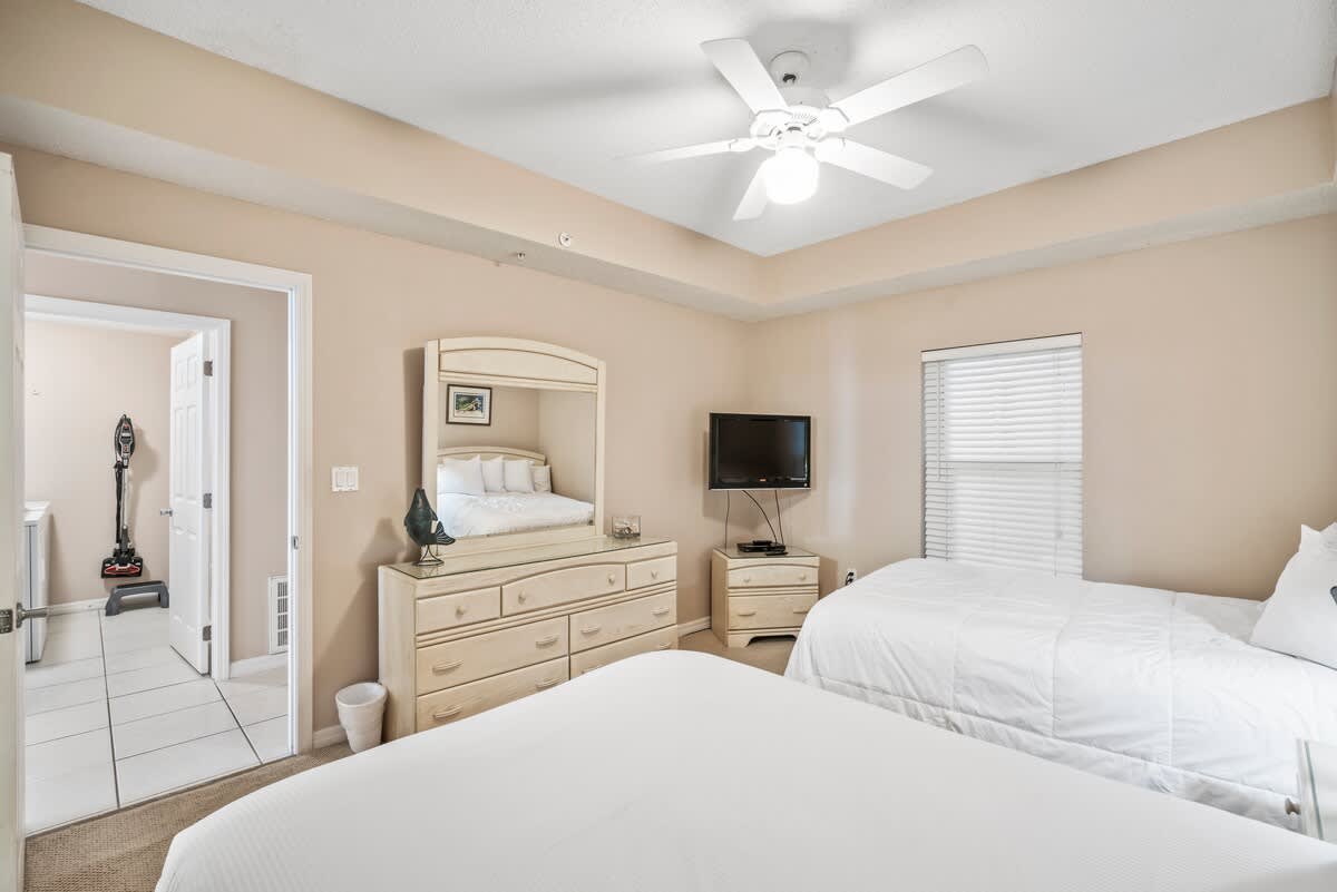 Second bedroom featuring two beds with white linens, a light-toned dresser with a flat-screen TV, and a window with blinds. A doorway leads to an adjacent tiled area.