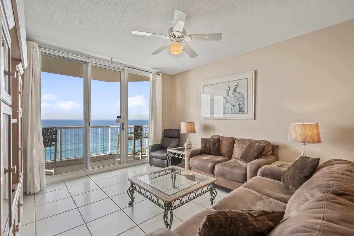 Inviting living room with two plush brown sofas, a glass coffee table, and a recliner, all facing sliding glass doors that lead to a balcony overlooking the ocean. A framed pelican print hangs on the wall.