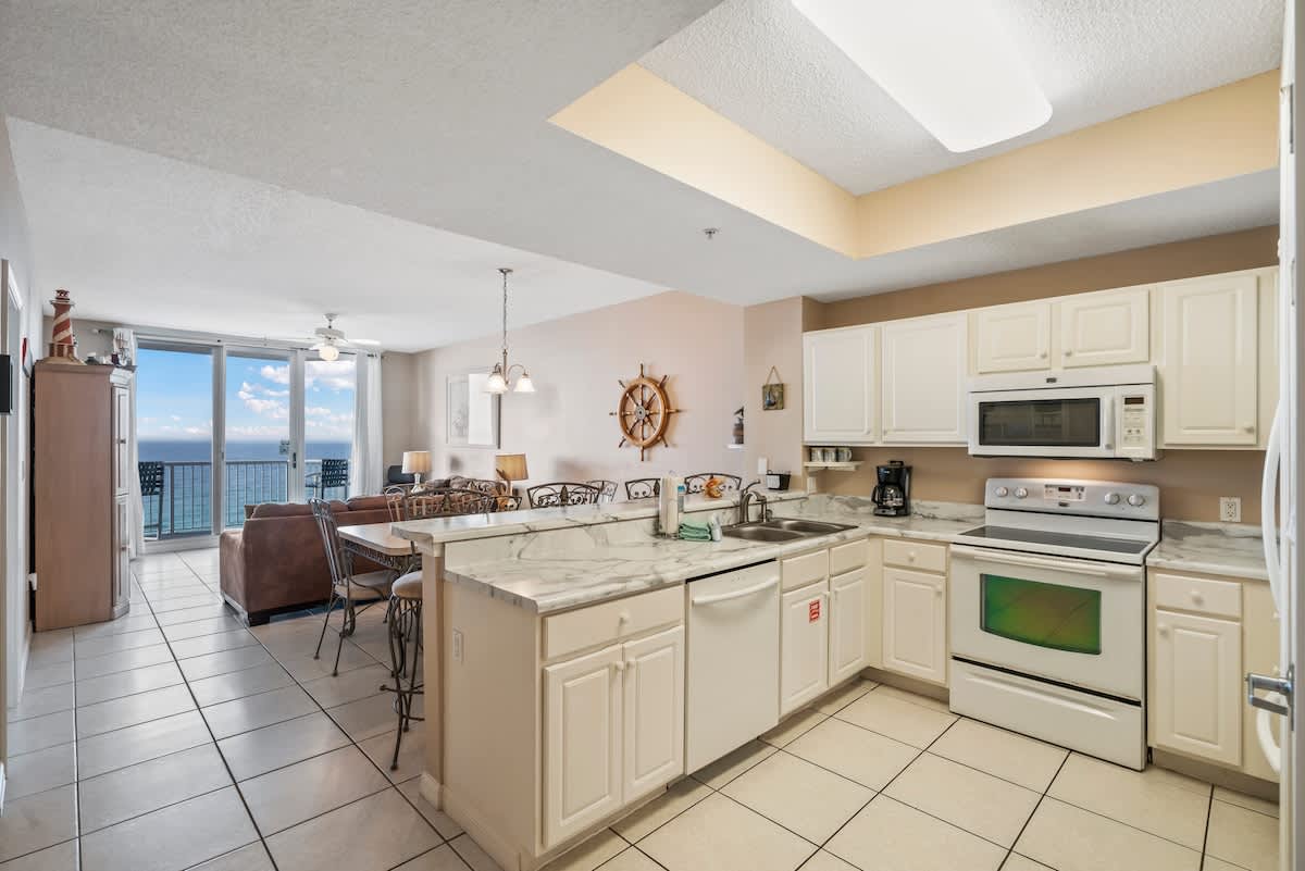 Expansive open-concept living area with a comfortable brown sofa, a dining table with wrought iron chairs, and a full kitchen with white cabinets and marble-look countertops. Sliding glass doors offer direct ocean views.