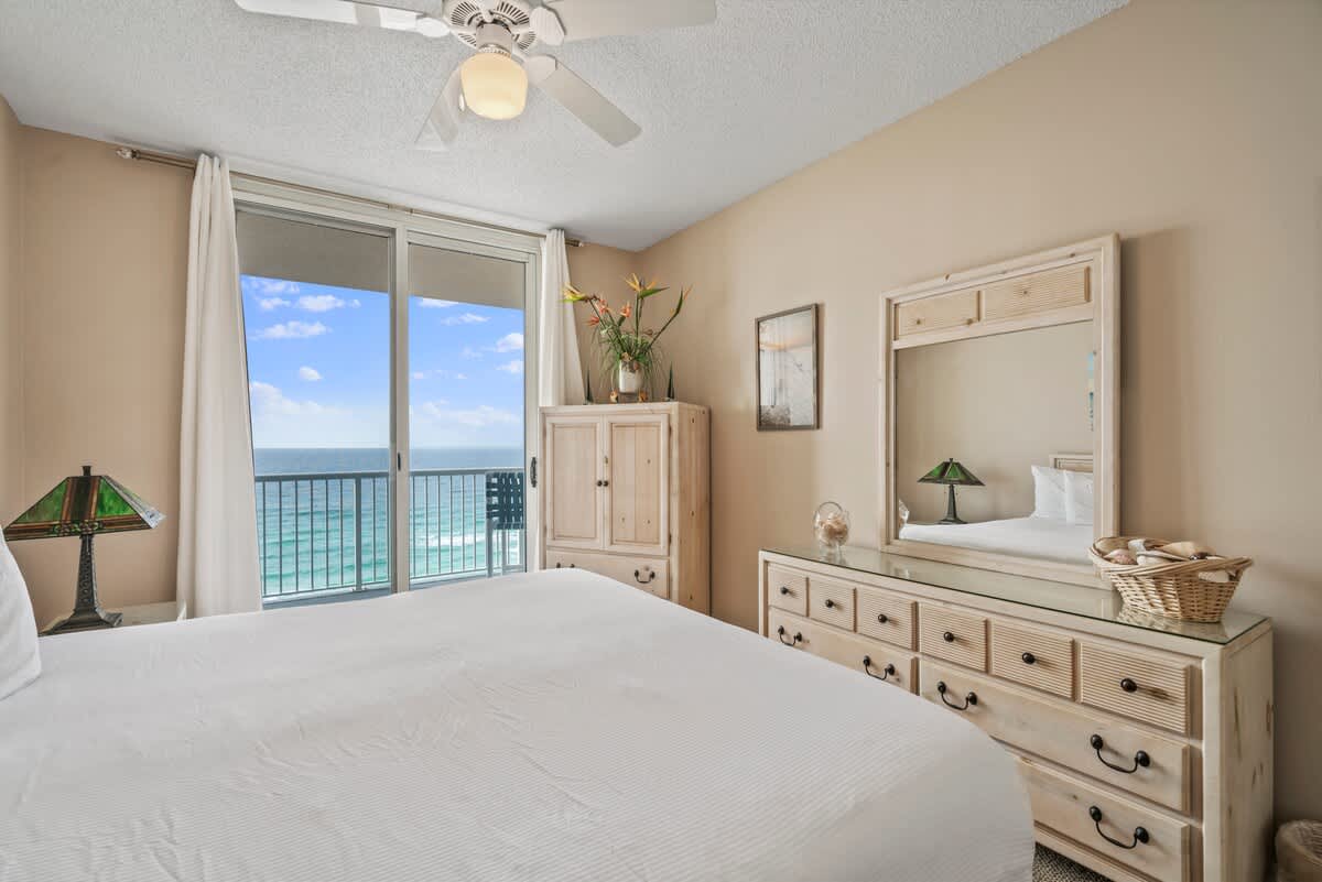 Master bedroom with a large bed, a light-toned dresser with a mirror, and a tall cabinet. Sliding glass doors offer panoramic ocean views and access to the balcony.