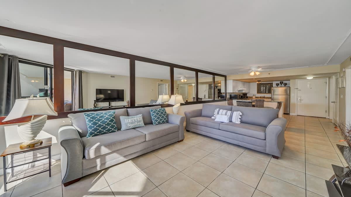 Bright living room featuring two comfortable gray sofas with decorative pillows, a side table with a lamp, and a large mirrored wall reflecting the space and a television.