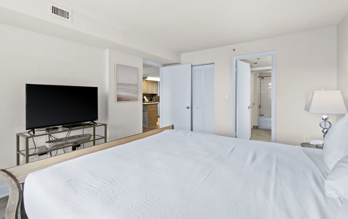 View of the king-sized bed with white bedding in bedroom-1, showing the flat-screen TV on a console and an open doorway leading to what appears to be a bathroom.