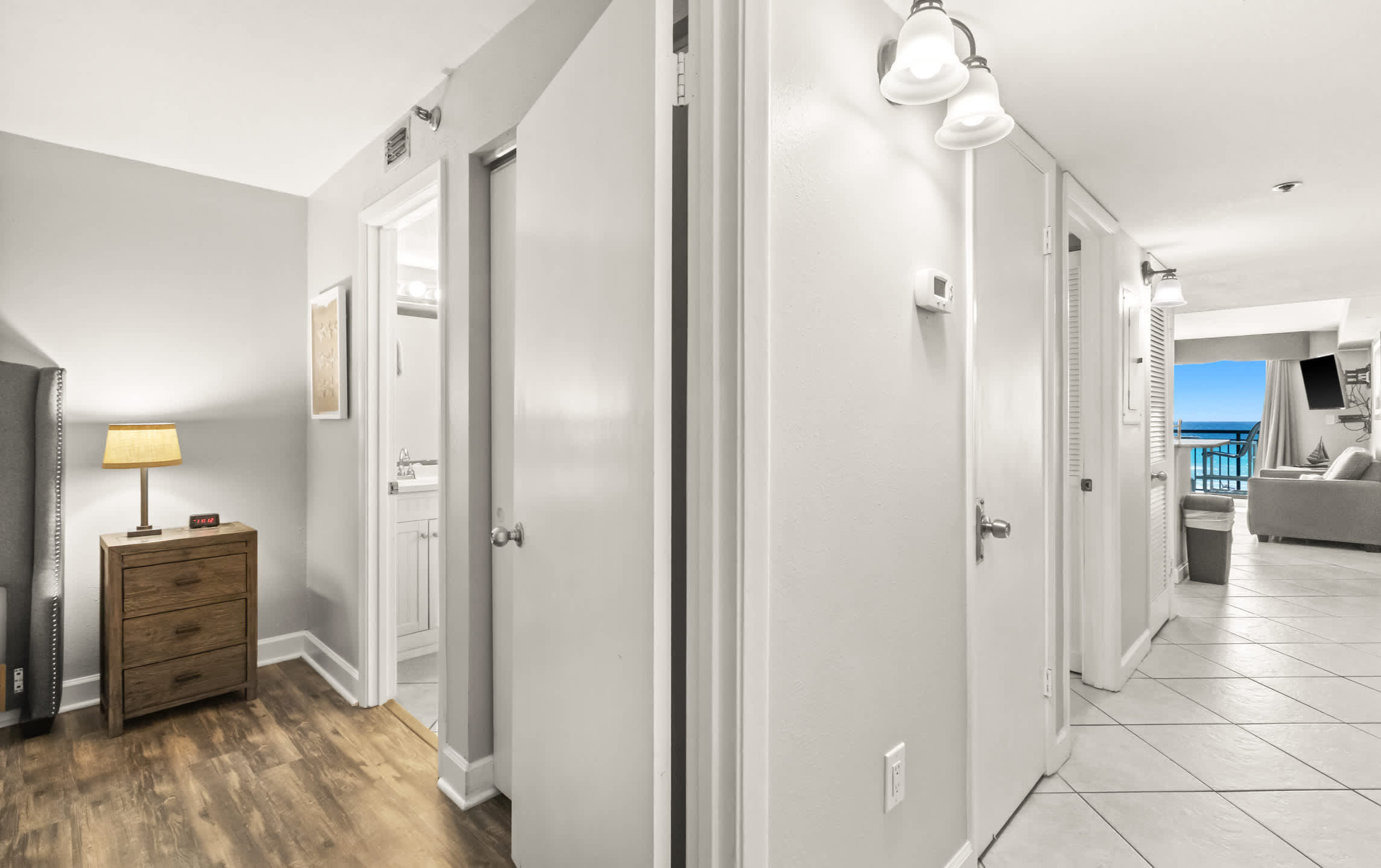 A bright internal hallway with light gray walls and wood-look flooring, featuring multiple white doors and wall sconces, offering a glimpse into a bedroom and a living area with an ocean view.
