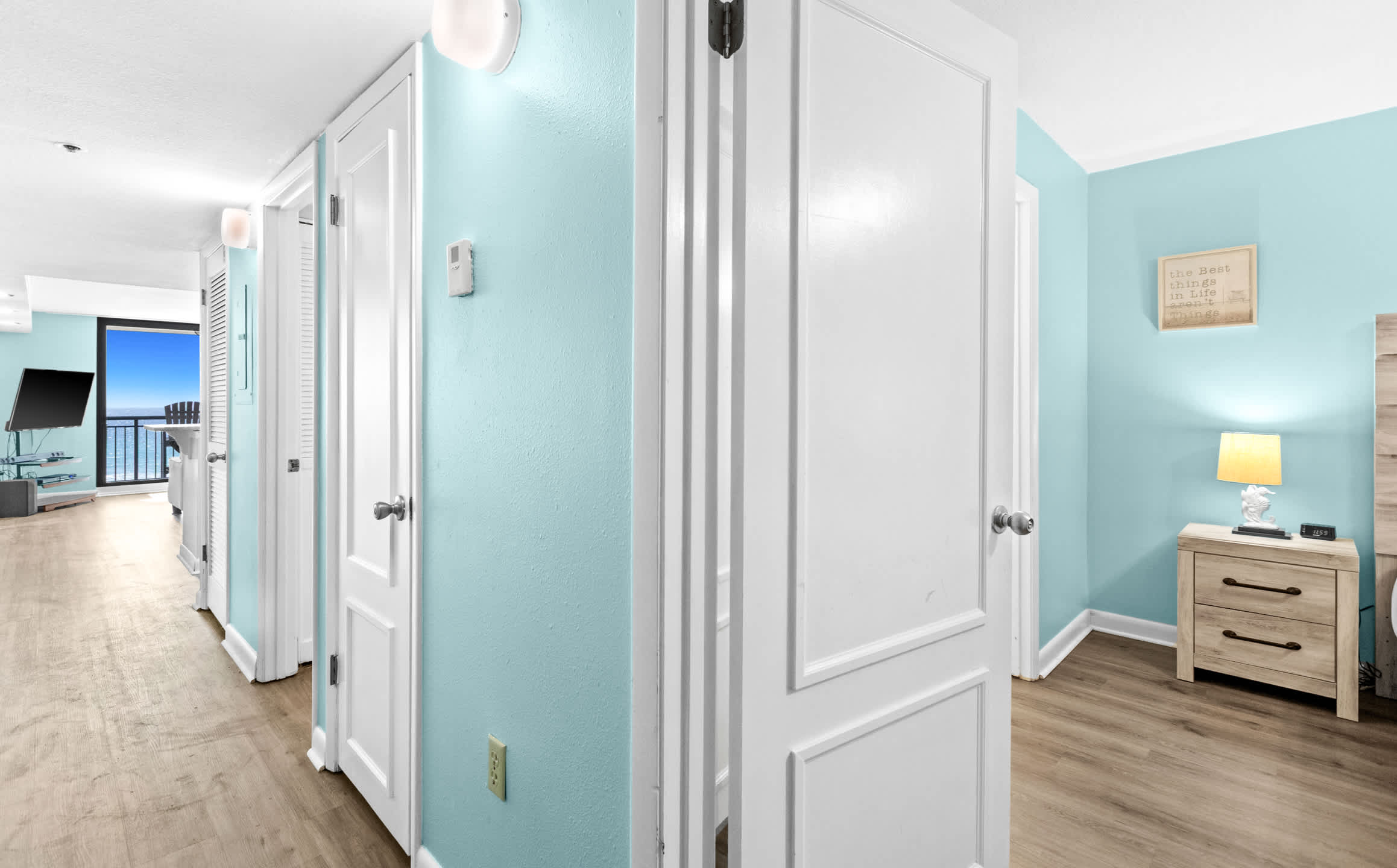A bright hallway with light blue walls and light wood-look flooring, featuring multiple white paneled doors leading to various rooms, with a glimpse of the living area and ocean beyond.
