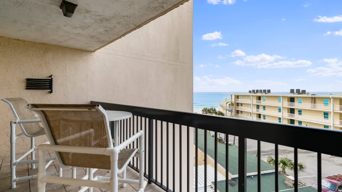 Outdoor balcony featuring two white bar-height chairs and a small round table, offering a partial ocean view and overlooking a multi-story building and parking lot below.