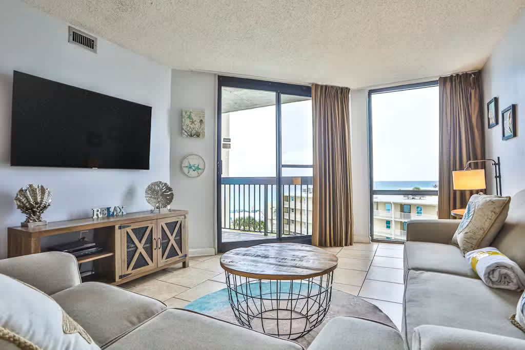 Bright living room with light gray walls, a large wall-mounted TV, a light-colored sofa, and a round wooden coffee table, featuring a balcony with ocean views through sliding glass doors.