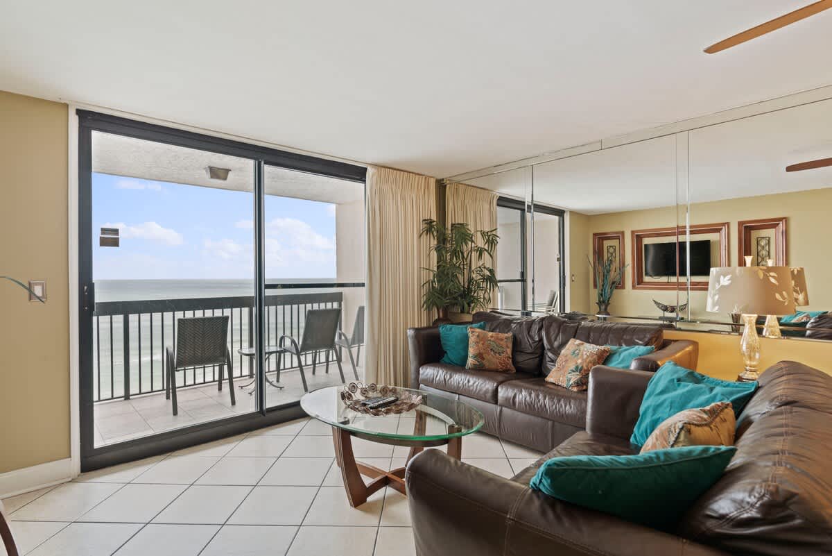 A spacious living room features two dark brown leather sofas with teal and patterned throw pillows, a glass-top coffee table, and large sliding glass doors opening to a balcony with ocean views. The room has light-colored tiled floors and a mirrored wall reflecting the space.