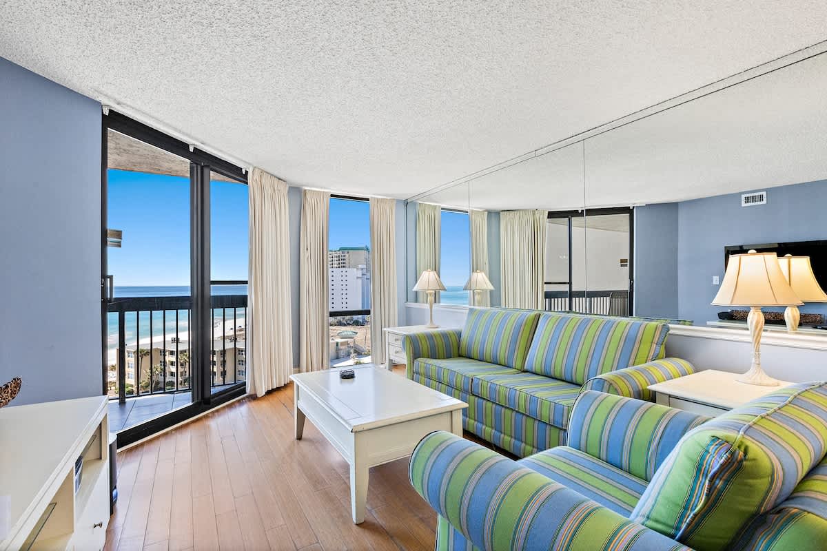 Bright living room with light blue walls, hardwood floors, and striped blue/green sofas, featuring a white coffee table and large sliding glass doors opening to a balcony with ocean views.