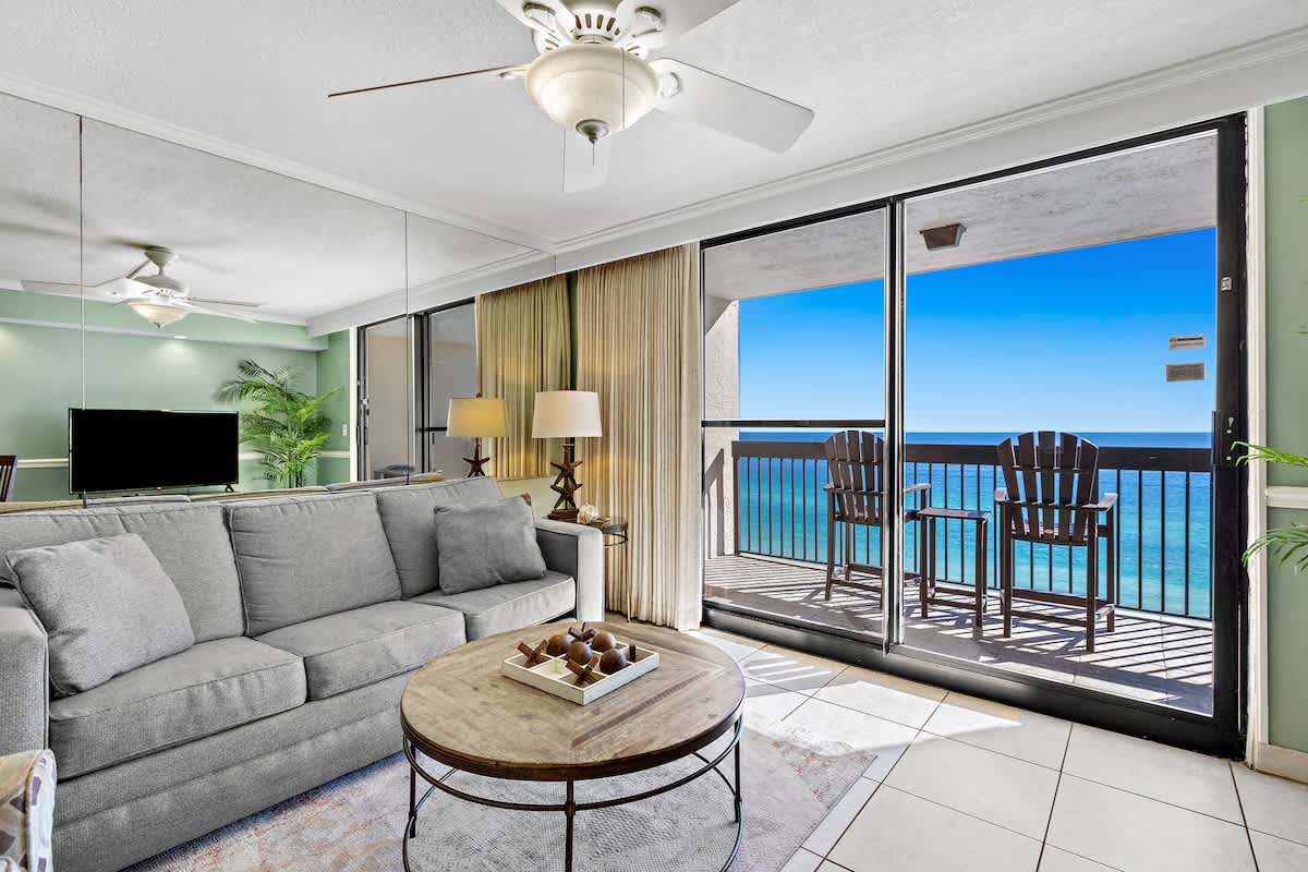 Bright living room with light green walls, a comfortable gray sofa, and a round wooden coffee table. A large mirror wall reflects the space and the stunning ocean view from the adjacent balcony.