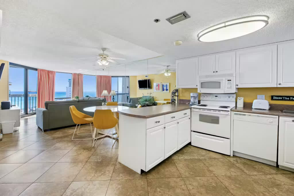 An open-concept living space features a kitchen on the right, a dining area with a round white table and yellow chairs, and a living room with ocean views through large windows. The floor is tiled, and a mirrored wall expands the space.