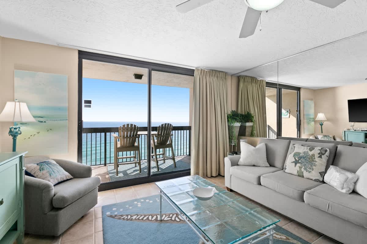 Bright living room with a light gray sofa, glass coffee table, and a blue rug with a starfish design, opening to a balcony with expansive ocean views through sliding glass doors.