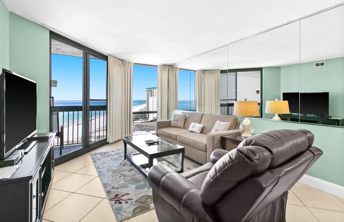 A bright living room with light green walls, a beige sofa, and a brown leather recliner, featuring large windows offering panoramic ocean and beach views. A dark wood TV stand and coffee table complete the space.