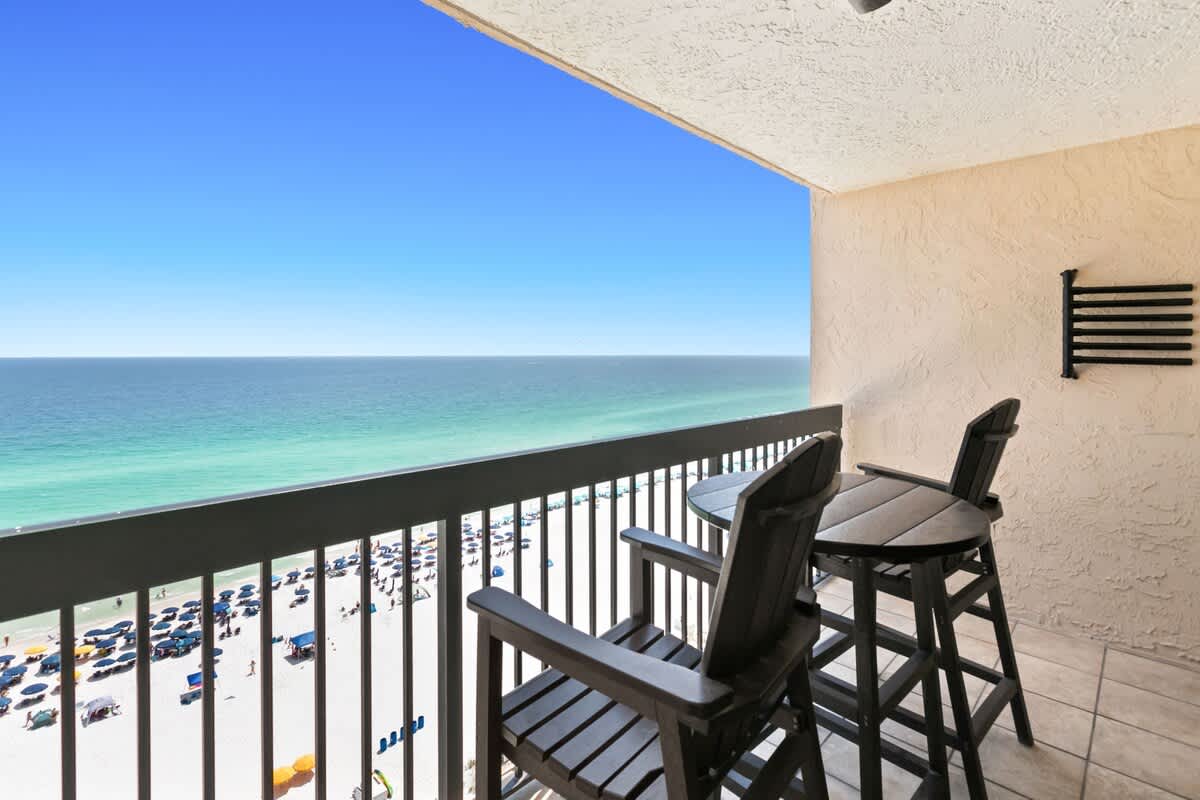An oceanfront balcony furnished with two dark wooden bar-height chairs and a matching table, offering expansive views of the clear blue ocean and the sandy beach below, dotted with umbrellas.