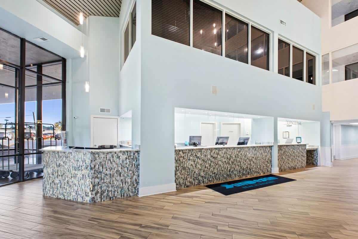 The resort's reception area features a long counter with a mosaic tile facade in shades of blue and brown, topped with a light surface. Multiple computer monitors are visible, and a "SunDestin Beach Resort" mat lies on the wood-look tile floor.