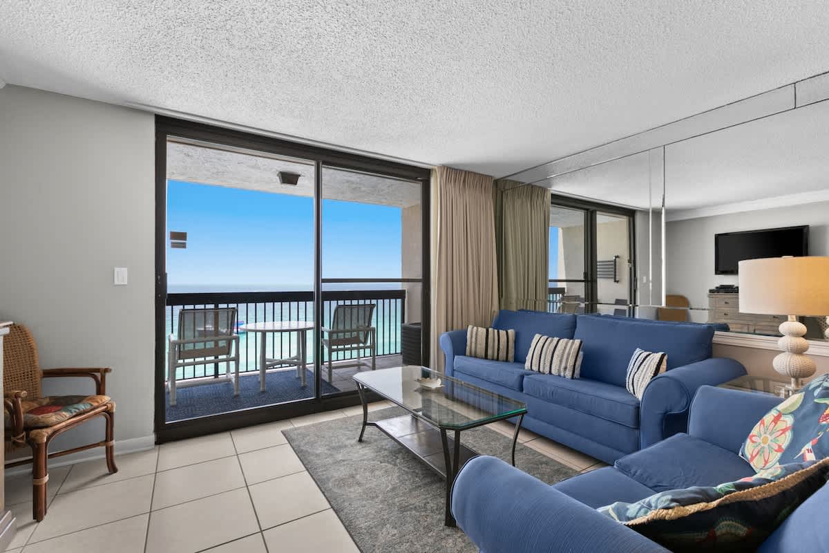 Bright living room featuring two blue sofas, a glass coffee table, and a large sliding glass door leading to a balcony with expansive ocean views. A mirrored wall reflects the room, enhancing the sense of space and light.