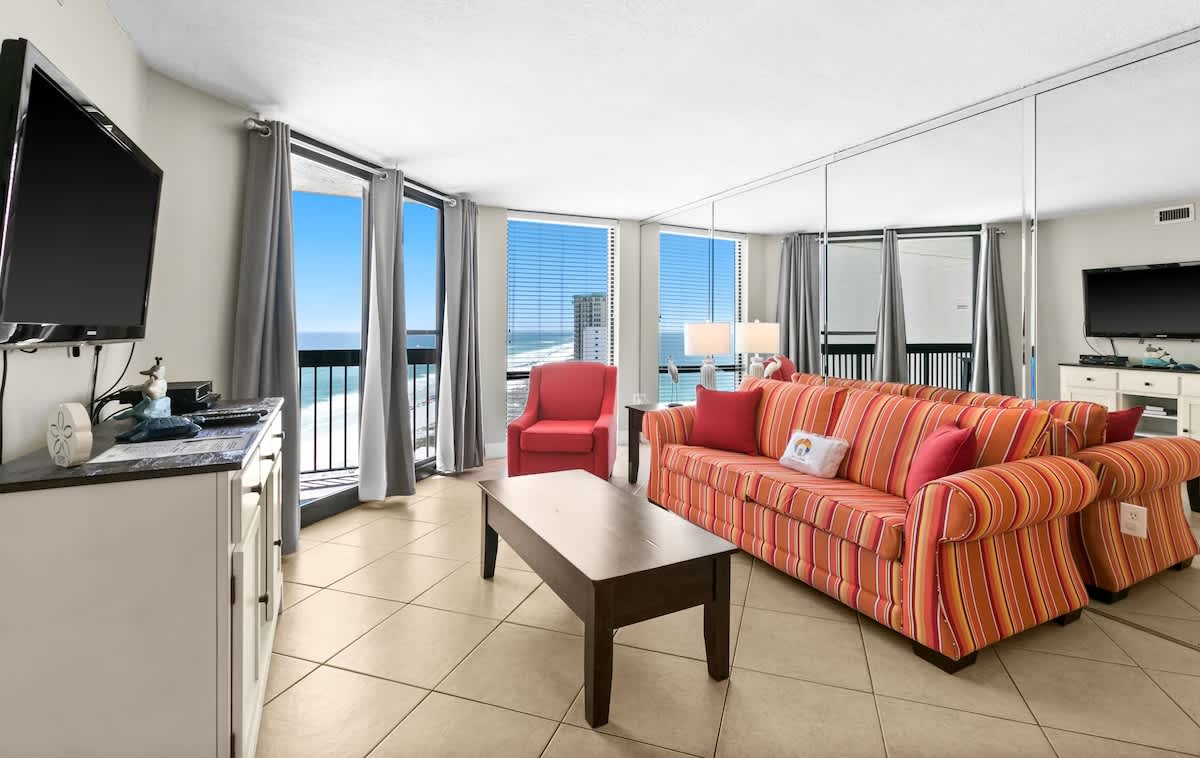 Bright living room with a striped orange sofa, red armchair, and dark wood coffee table on tiled floors. Large windows offer panoramic ocean views, with a flat-screen TV on a white console to the left.