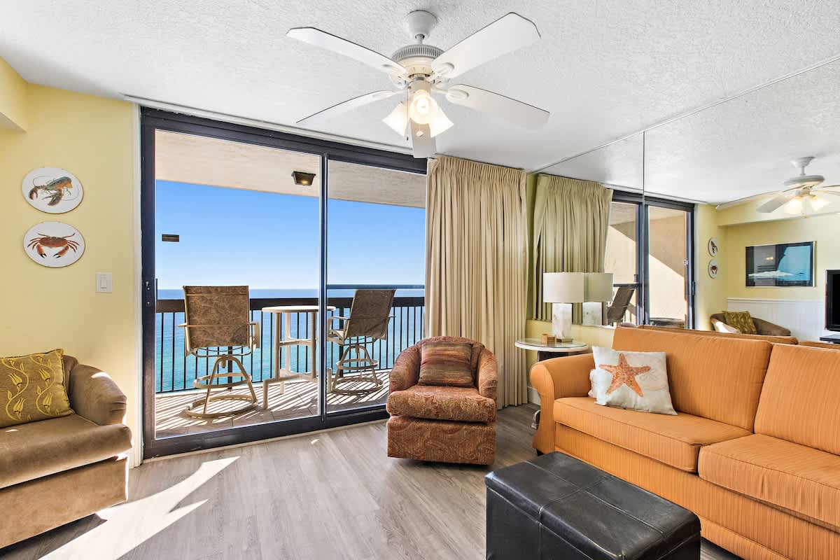 Bright living room with light yellow walls, a ceiling fan, and a large sliding glass door leading to a balcony with stunning ocean views. Features an orange sofa, a patterned armchair, and light wood-look flooring.
