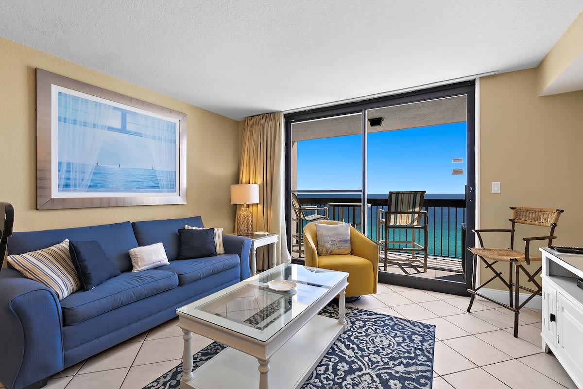 Bright living room featuring a blue sofa, a yellow armchair, and a glass-top coffee table on a patterned rug, with large sliding doors opening to a balcony overlooking the ocean.