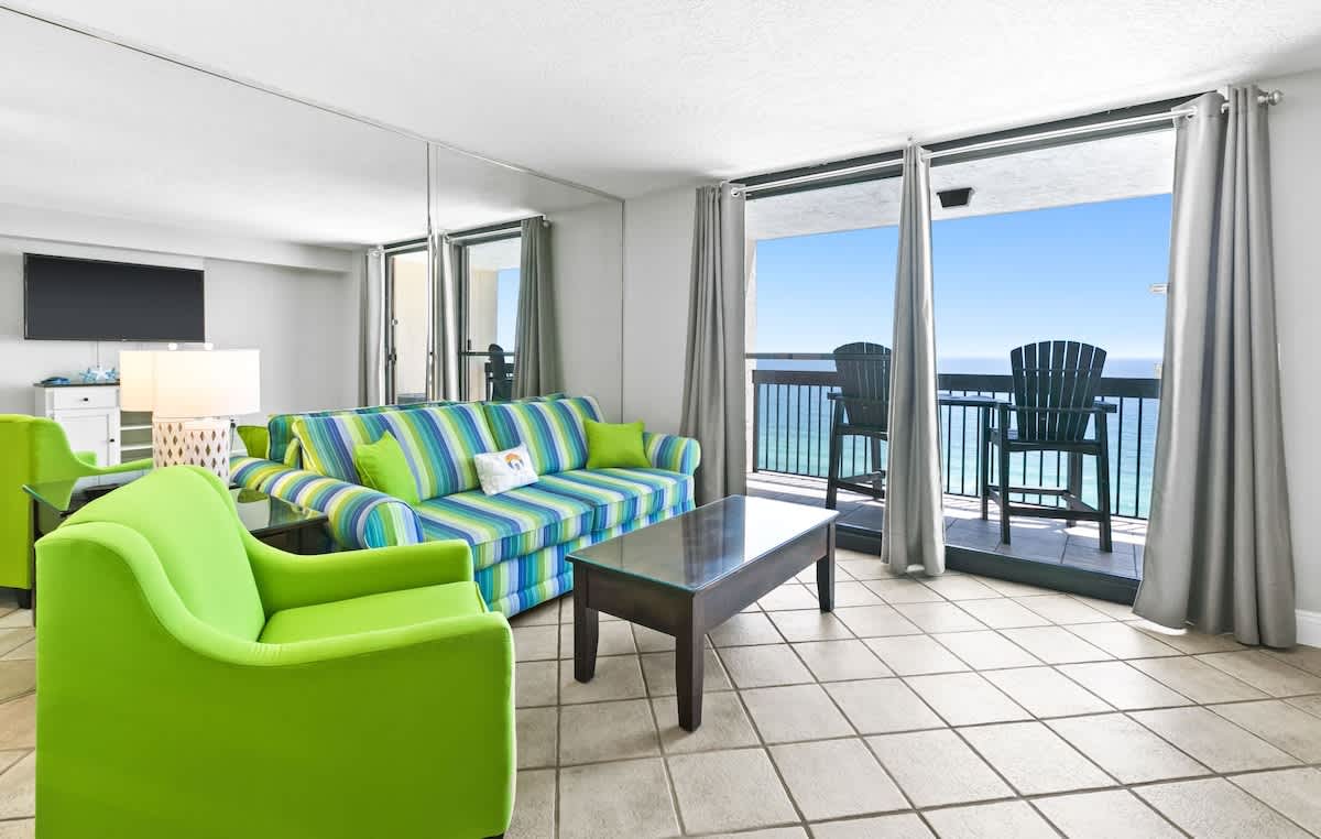 Bright living room with a vibrant striped blue and green sofa, lime green armchairs, a dark wood coffee table, and large sliding glass doors opening to a balcony with expansive ocean views.