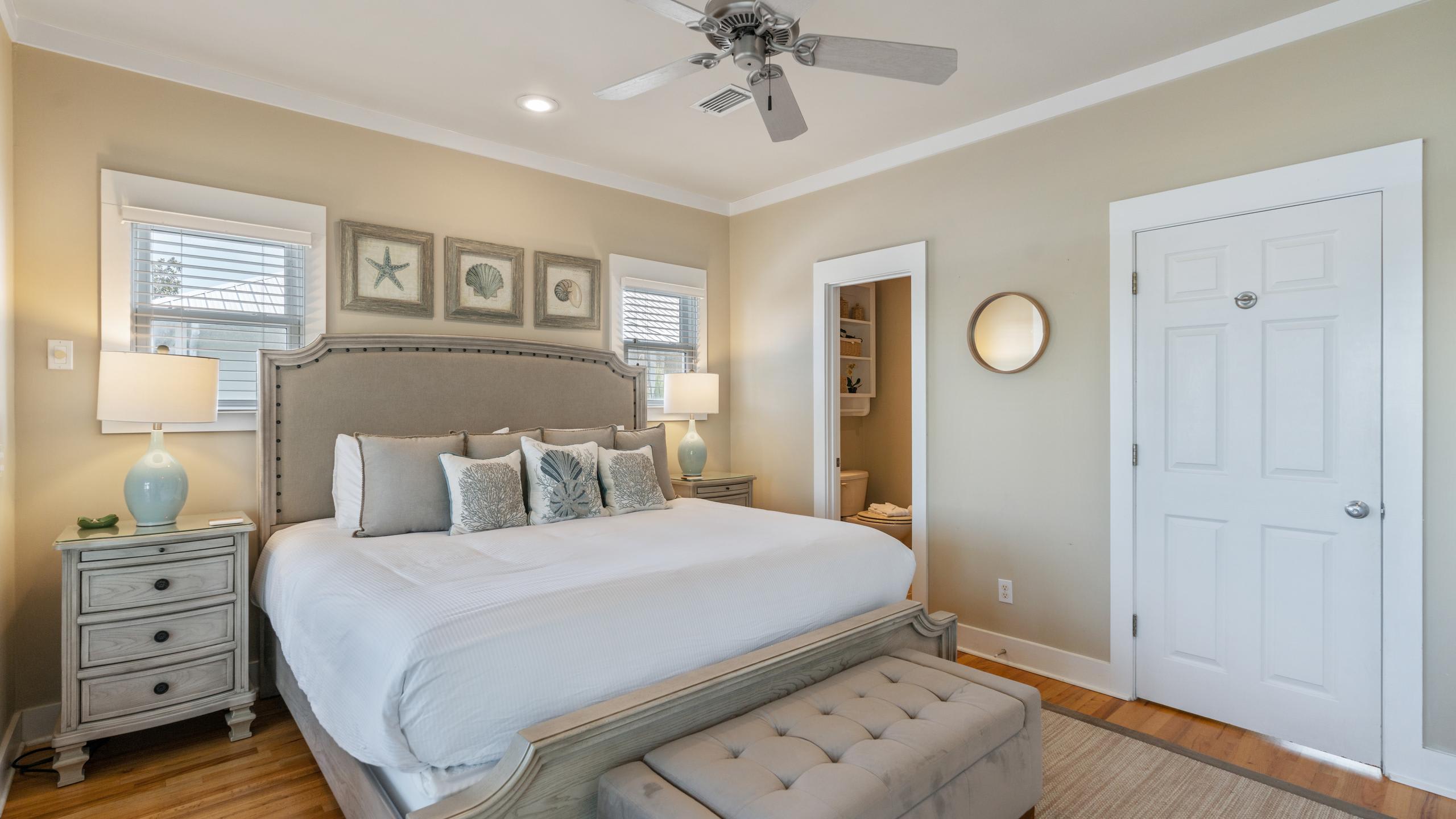 Elegant bedroom with a large king-sized bed, two nightstands with lamps, and multiple windows with white blinds. French doors lead to a balcony, and a round mirror is visible near a bathroom entrance.