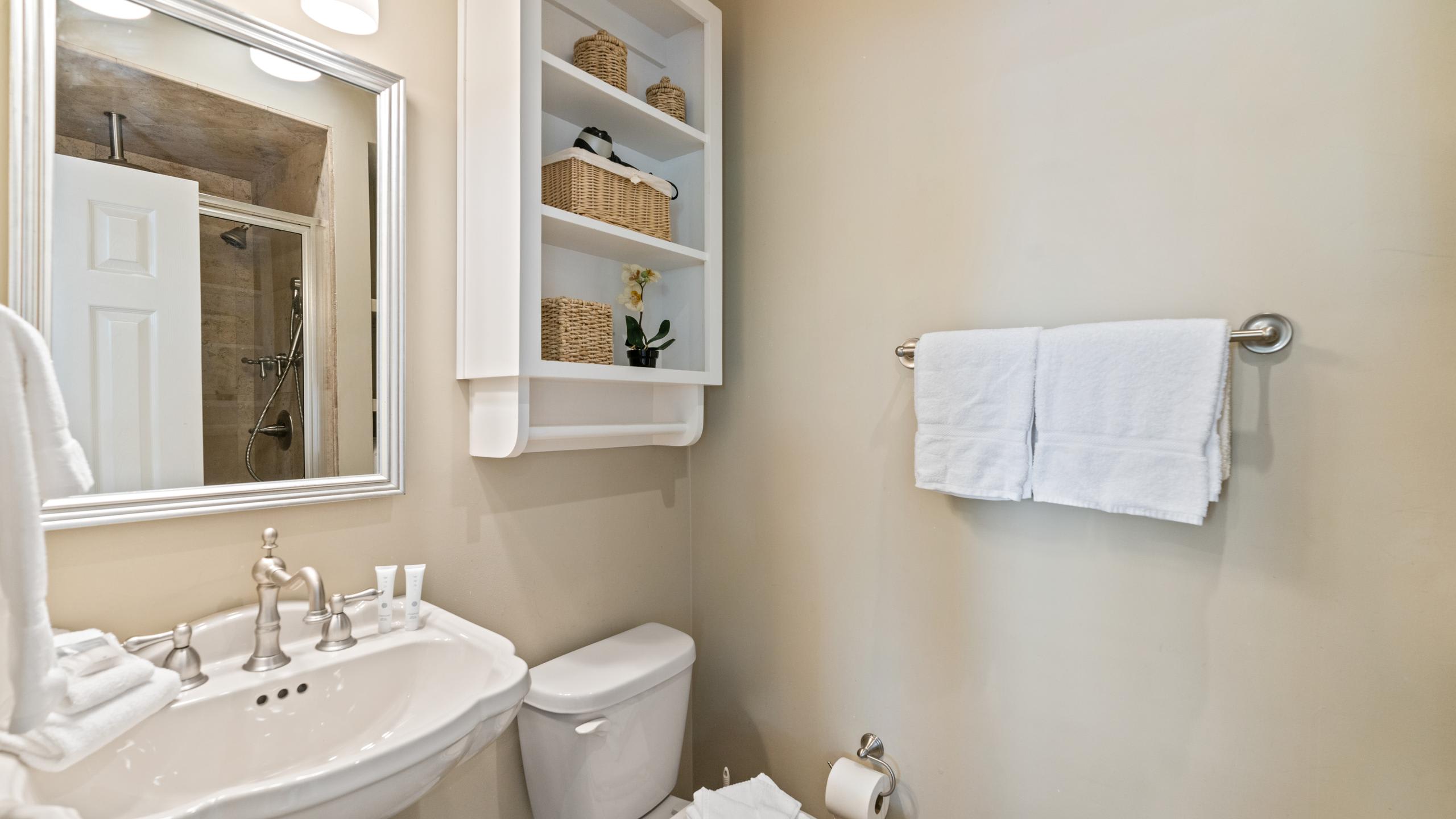 Bright bathroom with a white pedestal sink, a toilet, and a mirrored vanity. A white wall-mounted shelf with baskets and a towel rack with white towels are visible.