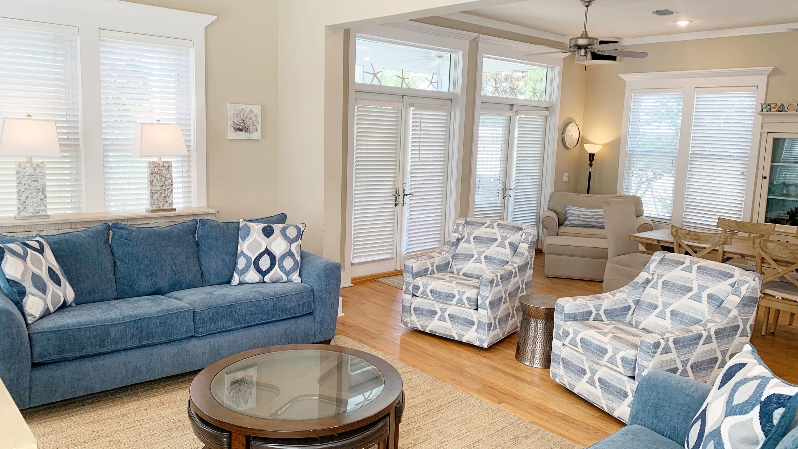 A bright living room features a blue sofa with patterned pillows, two patterned armchairs, and a round wooden coffee table on a textured rug. White blinds cover large windows, and a ceiling fan hangs above.