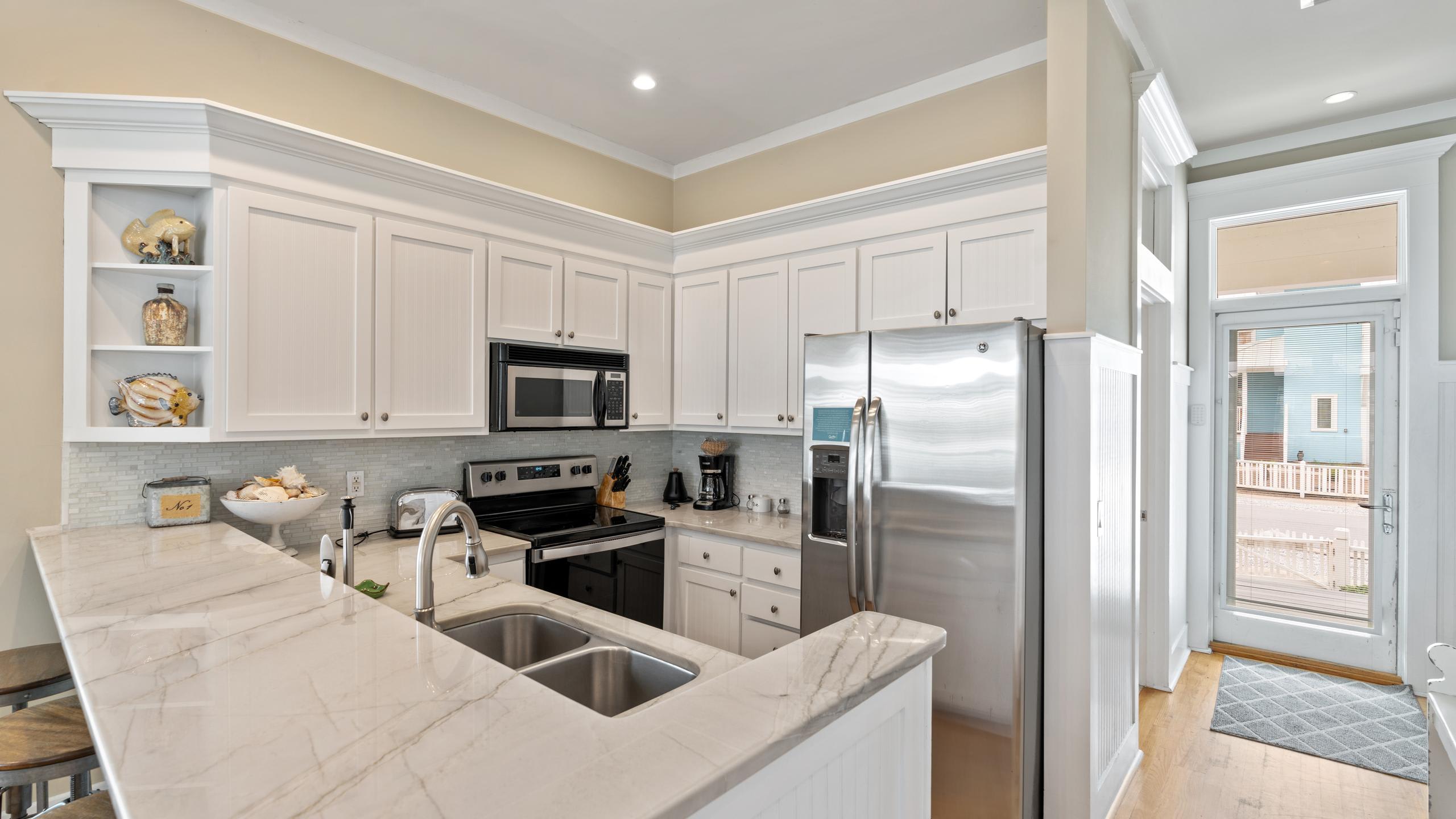 A clean kitchen with white cabinets and marble countertops, featuring a stainless steel refrigerator and microwave. A double sink is visible, and the space opens up to an entryway with a view of another house.