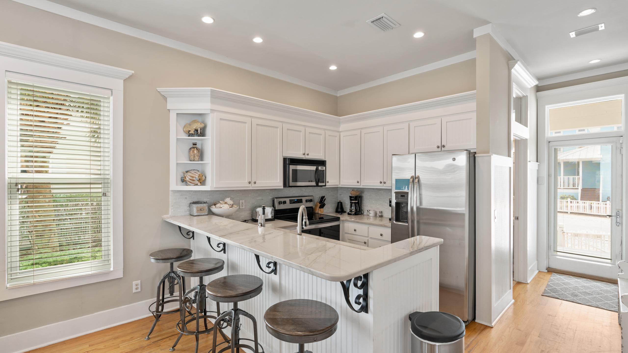 A spacious kitchen with white cabinets, marble countertops, and a breakfast bar with three wooden bar stools. Stainless steel appliances are integrated, and the room features recessed lighting and hardwood floors.