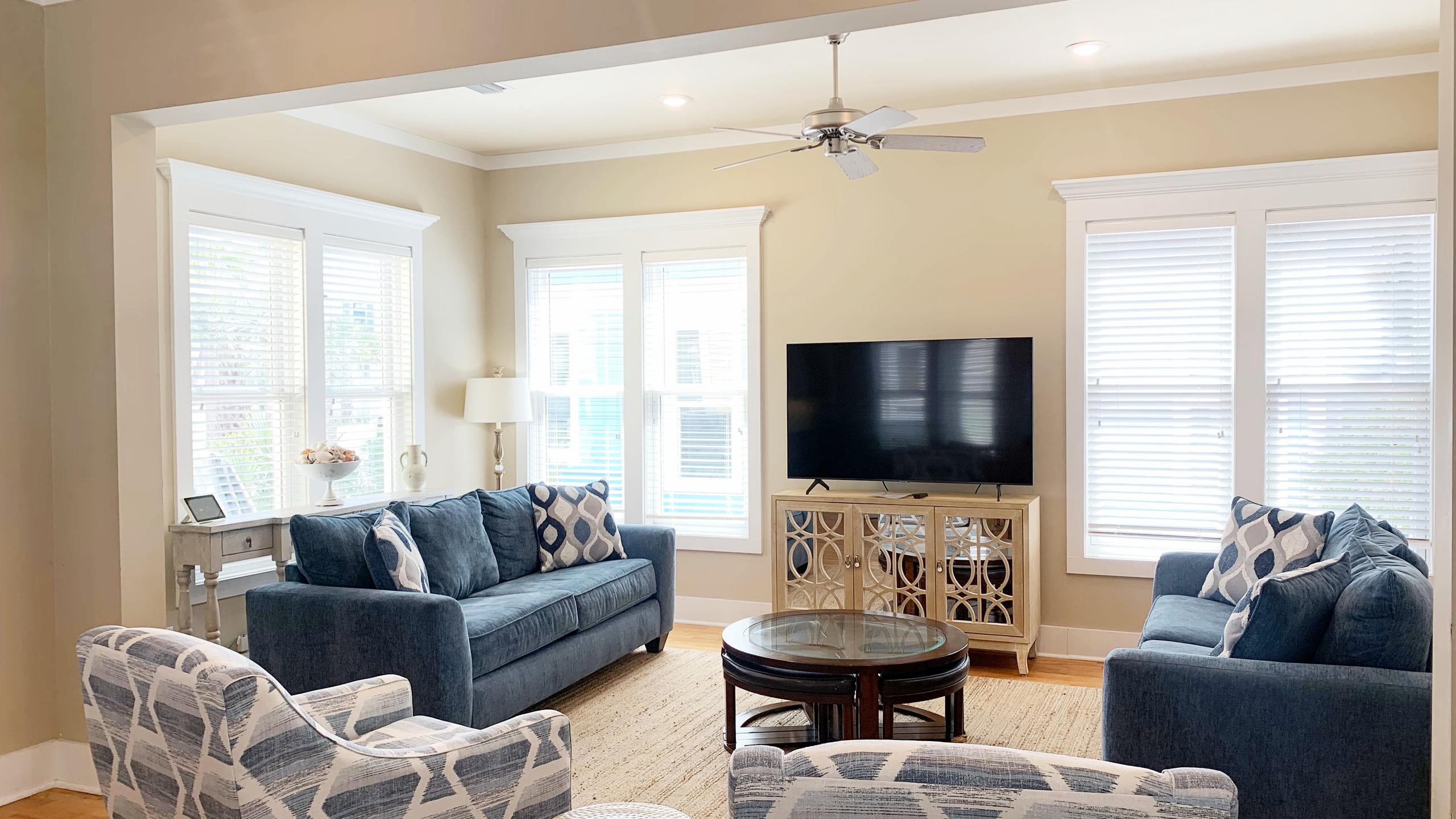 A bright living room with light beige walls features two blue sofas with patterned throw pillows, a large flat-screen TV on a light wood console, and a round dark wood coffee table on a natural fiber rug.